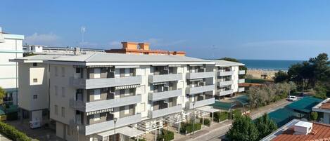 A multi-story residential building with balconies, a parking area, and a view of the sea.