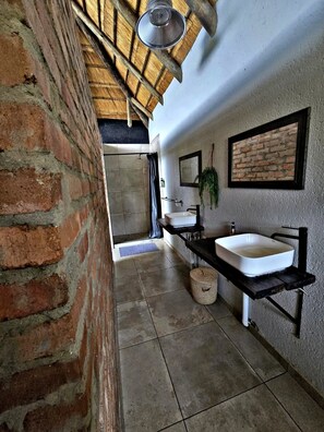 Un baño con pared de ladrillo, techo de madera, lavamanos blanco, espejo y planta colgante.