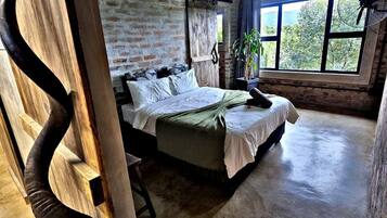 Ein rustikales Schlafzimmer mit Backsteinwand, Holzbalkendecke, einem Bett mit weißen Leinentüchern, einer Pflanze und einem Fenster mit Blick auf Bäume.