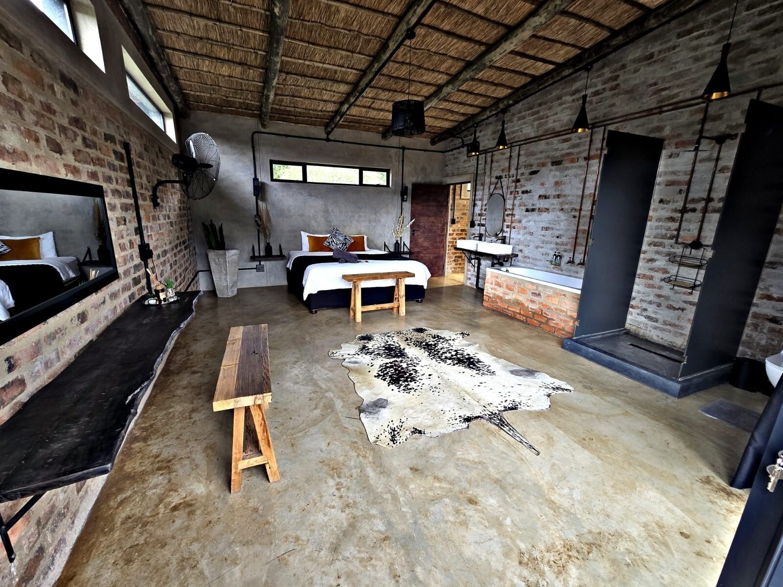 A rustic room with exposed brick walls, a bed with a headboard, a wooden bench, a cowhide rug, and a hanging light fixture.