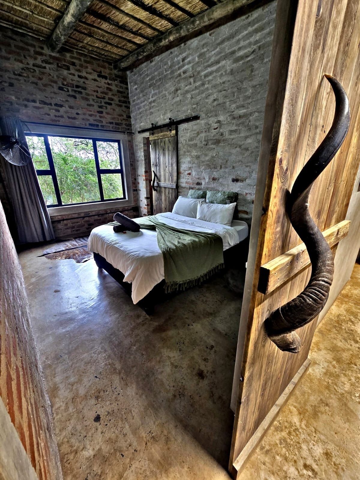 A rustic room with a bed, a wooden door with a curved handle, and a window with a view of trees.
