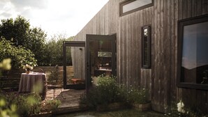 A modern wooden building with an open patio area, a table with chairs, and a small garden. - Unique Cabin Wrapped in Beautiful Welsh Scenery (Presteigne)