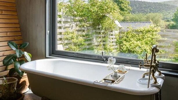 A modern bathroom with a freestanding bathtub, a large window offering a view of greenery and buildings, and a wooden ceiling. - Unique Cabin Wrapped in Beautiful Welsh Scenery (Presteigne)