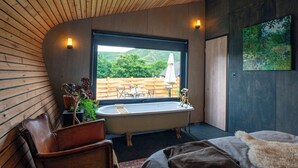 A modern bathroom with a freestanding bathtub, wooden ceiling, and a large window overlooking a garden. - Unique Cabin Wrapped in Beautiful Welsh Scenery (Presteigne)
