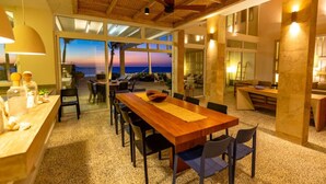 Un comedor espacioso con una larga mesa de madera, sillas y vistas al océano al atardecer.