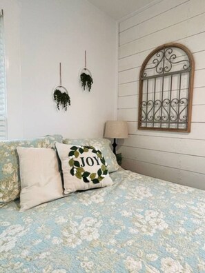 Une chambre avec une couvre-lit à motifs floraux, un coussin orné du mot « JOIE », une fenêtre avec des barreaux en fer décoratifs et deux plantes suspendues.