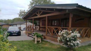 A wooden cabin with a porch, a car parked in front, and a garden with a statue and flowers. - CHALET L'AVENTURE 14km from VANNES (Sulniac)
