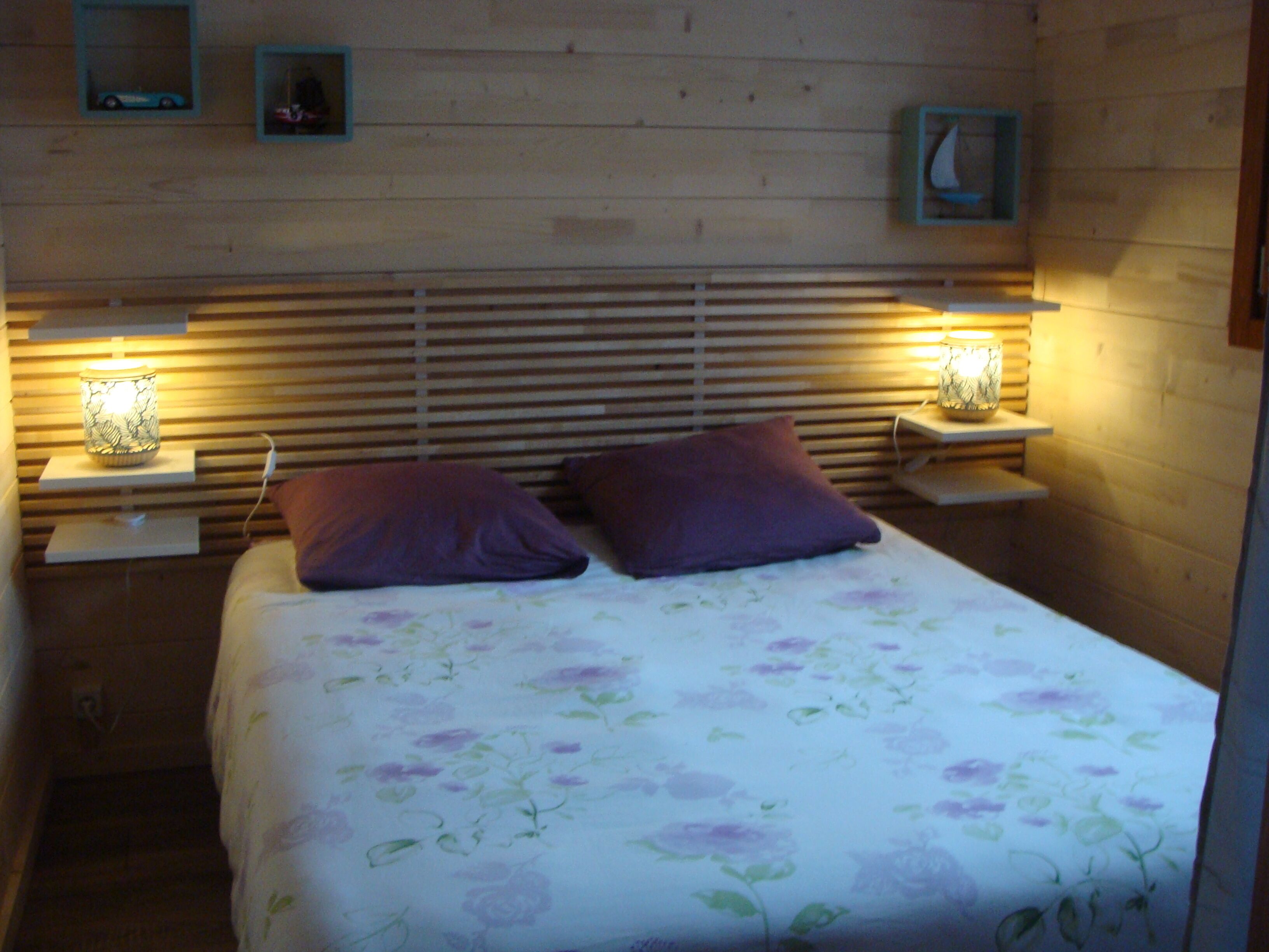 Un dormitorio con cabecera de madera, dos lámparas de noche, colcha estampada con motivos florales y dos almohadas moradas.
