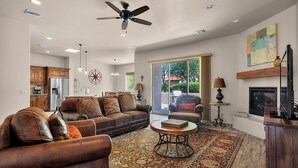 A spacious living room with a large brown leather sofa, a coffee table, and a fireplace. There is a ceiling fan, recessed lighting, and a view of the outdoors through large windows. - Casita Paz at The Tubac Sanctuary (Tubac)