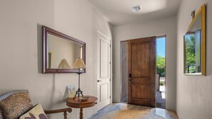 A hallway with a wooden door, a round rug, a chair with a cushion, a small table with a lamp, and a mirror on the wall. - Casita Paz at The Tubac Sanctuary (Tubac)