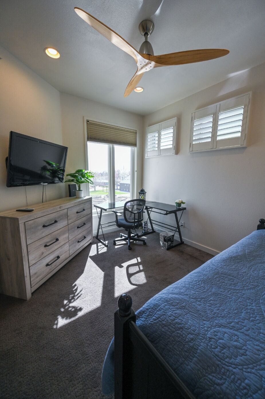 A bedroom with a bed, a desk, a chair, a TV, a ceiling fan, and a window with blinds.
