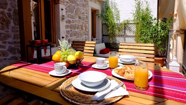 Una mesa servida con platos, un frutero y vasos de jugo de naranja, con una pared de piedra y muebles de madera al fondo.
