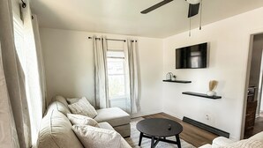 A modern living room with a sofa set, a round coffee table, a ceiling fan, a flat-screen TV, and a small shelf with decorative items.