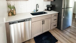 A modern kitchen with light wood cabinets, a stainless steel dishwasher, a black refrigerator, and a white tiled backsplash.