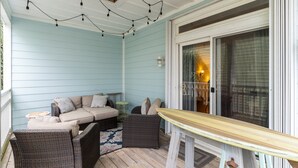 A covered porch with a beige sofa, wicker chairs, and a wooden table. - Steps to the Sand in Carolina Beach, Sleeps 10! Villa (Carolina Beach)
