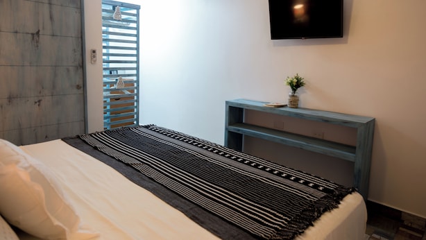 Une chambre à coucher avec un lit, un téléviseur fixé au mur, une petite table avec un vase et une fenêtre avec des stores.