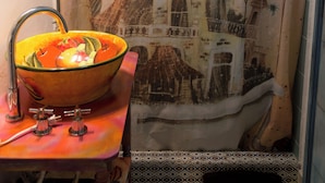 Une salle de bain avec un rideau de douche représentant un bâtiment de style colonial, un lavabo avec un receveur et un tapis à motifs à l’entrée.
