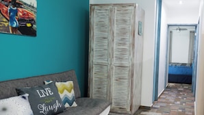 Un salon aux murs de couleur turquoise, avec un canapé gris orné de coussins à motifs, une armoire en bois et un couloir au sol à motifs menant à une chambre.