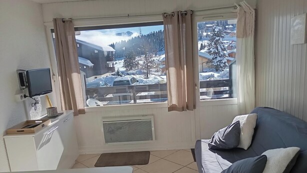 A cozy living room with a large window, a flat-screen TV mounted on the wall, a sofa with cushions, and a small table. - 5 minutes from the La Legette chairlift, Studio-Cabin 21m² Les Saisies (Hauteluce)