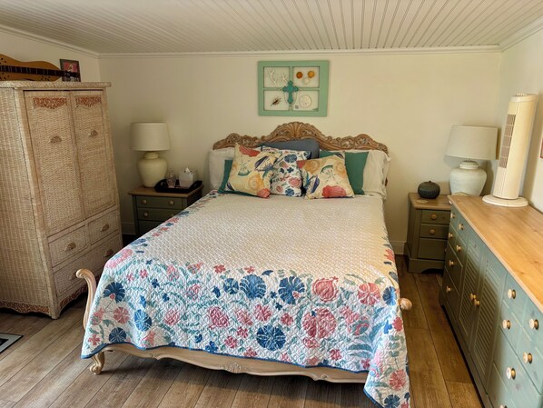 A bedroom with a floral quilted bedspread, a wooden headboard, two bedside tables with lamps, a dresser, and a wall clock.