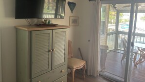 Een kamer met een aan de muur bevestigde televisie, een kast met lades, een stoel en uitzicht op een terras door een schuifdeur.