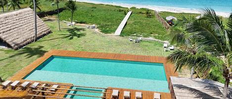 Ein Poolbereich mit Holzterrasse und Liegestühlen, umgeben von einer Rasenfläche und Palmen, mit Blick auf einen Strand.