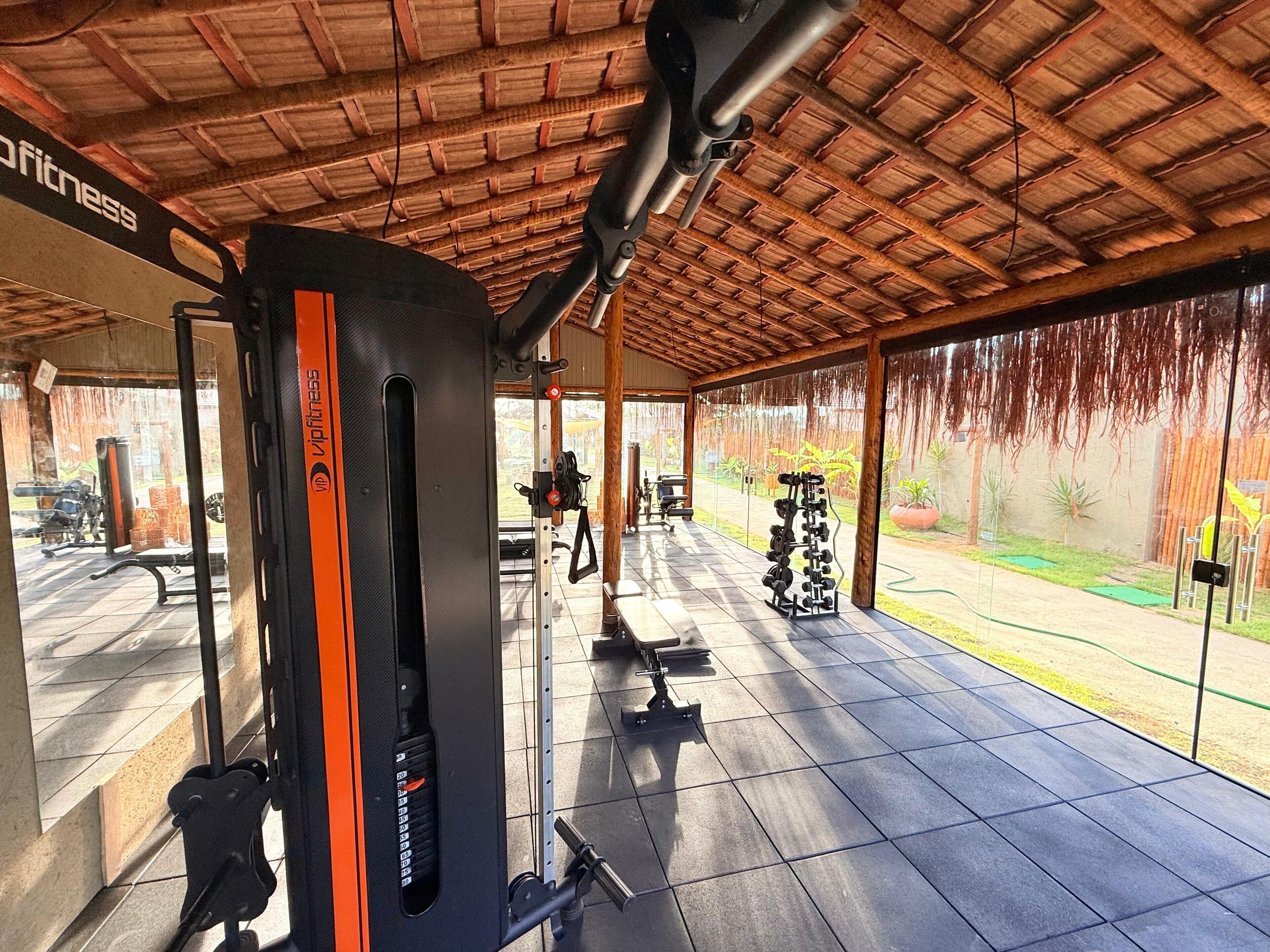 A covered outdoor gym area with exercise equipment, including a bench press and dumbbells, under a wooden roof.