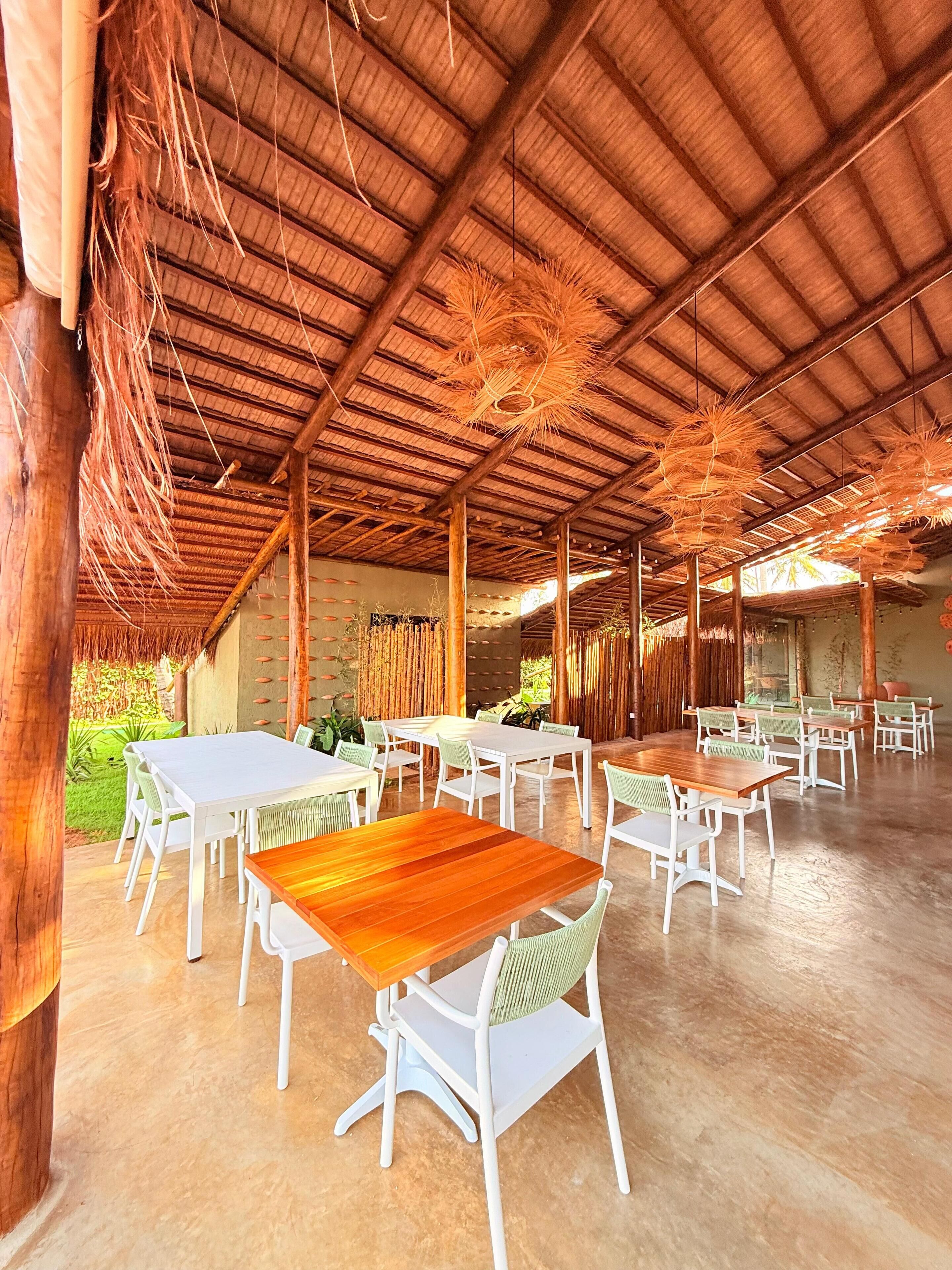 Een openluchtdinergedeelte met meerdere tafels en stoelen, een hoog plafond met houten constructie en uitzicht op groen.