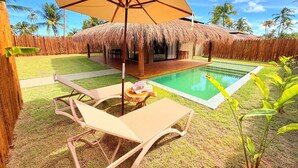 Un área junto a la piscina con una silla de descanso, una mesa con una bebida y una estructura de techo de paja.