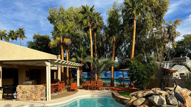Zona de piscina con área de estar cubierta, rodeada de palmeras y vista al océano.