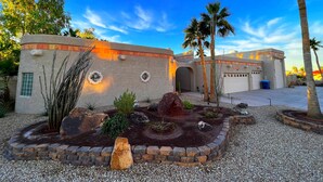Edificio de estilo desértico con fachada curva, un pequeño jardín con rocas y cactus, y palmeras.