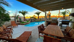 Zona junto a la piscina con muebles de madera, una barbacoa y vistas al atardecer.