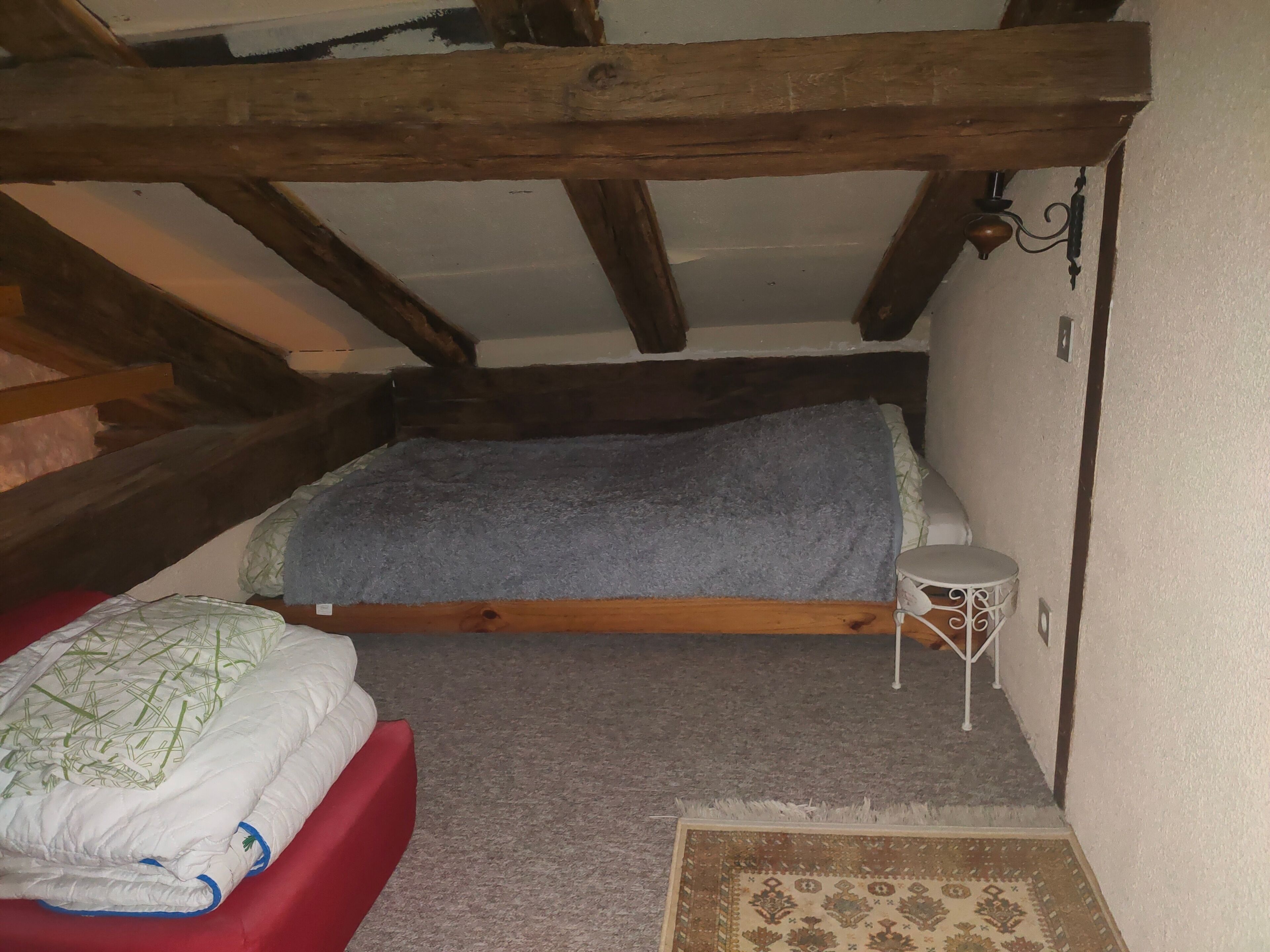 A small, single-bed room with a sloped ceiling, a red bed, a small bedside table, and a patterned rug.