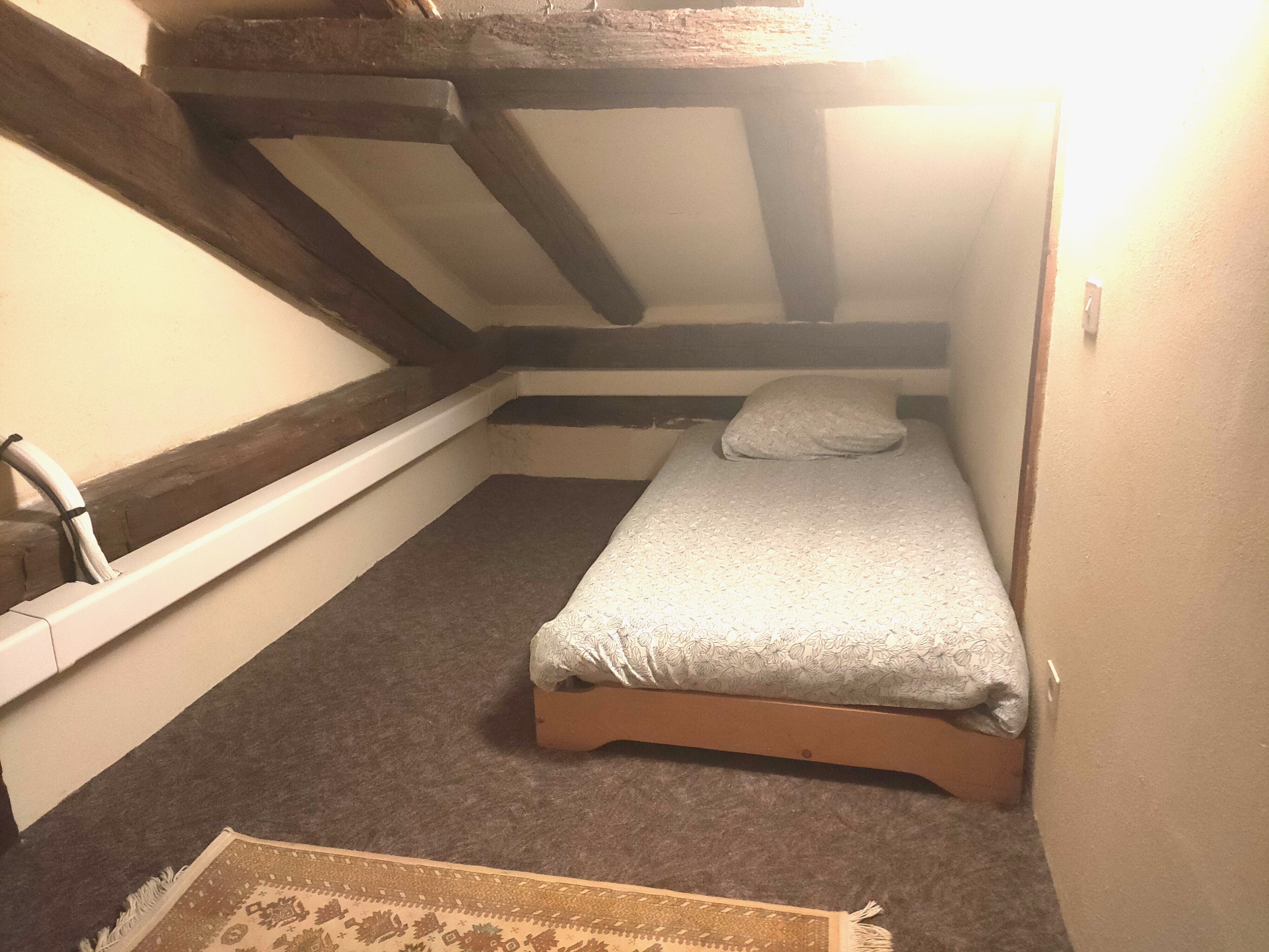 Una stanza piccola e stretta con un letto singolo, una finestra e un soffitto con travi in legno.