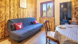 A cozy room with a blue sofa, a wooden wall, a window, a table with a patterned tablecloth, and a refrigerator. - Le Grand Som - Proche Station de ski (Saint-Pierre-de-Chartreuse)