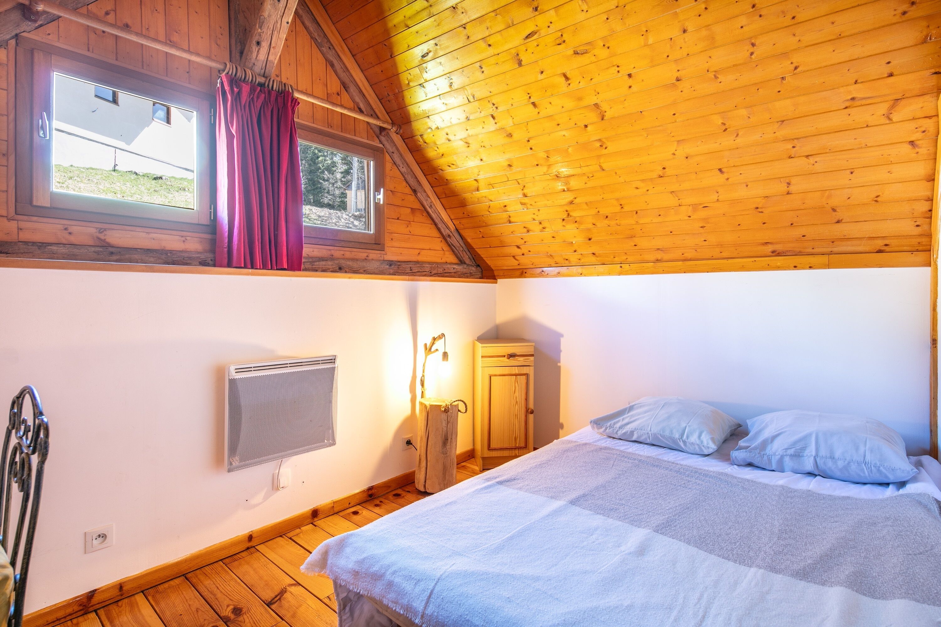Une chambre avec un plafond en bois, un lit avec des oreillers, un radiateur et une fenêtre avec des rideaux.