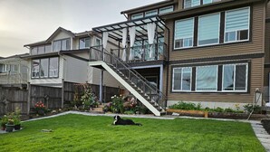 A two-story house with a covered outdoor area and a dog lying on the grass. - Private south-facing suite with a full bathroom and fully equipped kitchen. (Coquitlam)