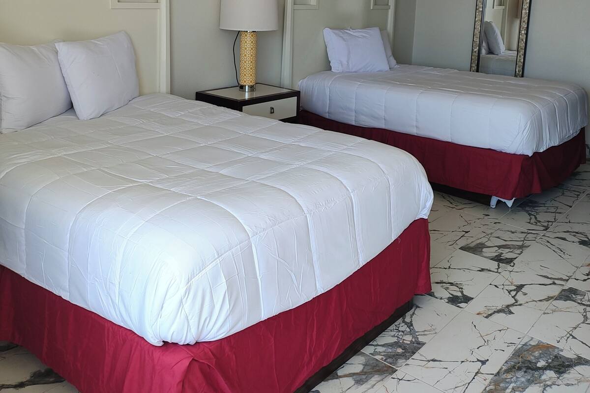 A hotel room with two beds, a nightstand with a lamp, a mirror, and marble-patterned flooring.