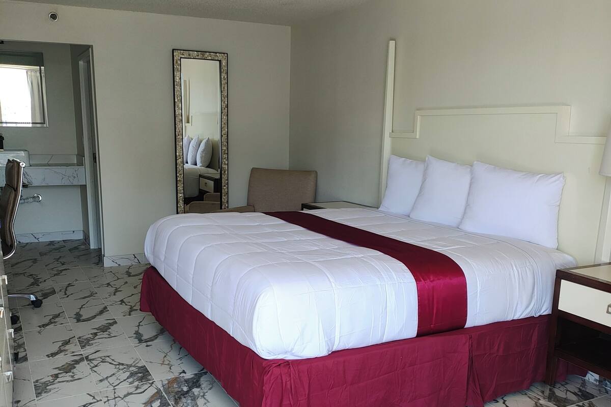 A hotel room with a large bed, a desk, a chair, a mirror, and a bathroom visible through an open door.