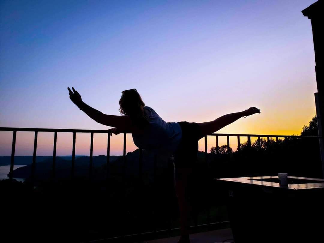 A person standing with arms outstretched on a balcony overlooking a body of water during sunset.