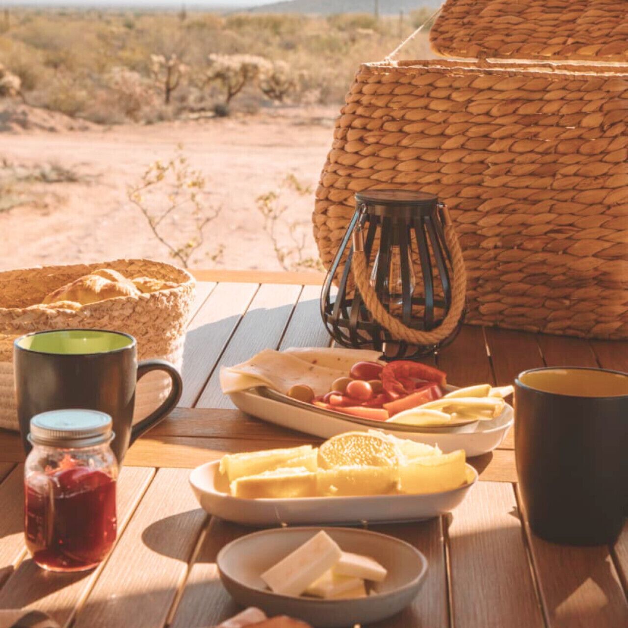 A wooden table with a woven basket, a metal lantern, a plate of food, a jar of jam, and two mugs.