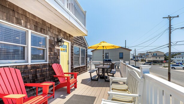 A wooden deck with red Adirondack chairs, a yellow umbrella, and a white railing overlooking a street.