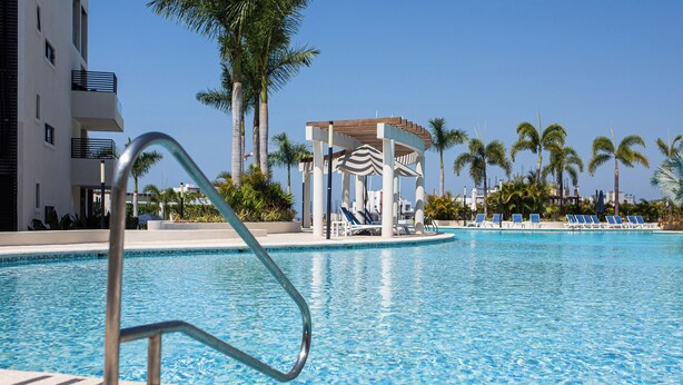 A swimming pool with lounge chairs, a gazebo, palm trees, and a modern building with balconies. - 3 bedroom penthouse in Bucerias - Ocean view, BBQ and hotel-like amenities (Bucerías)