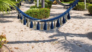A hammock in a sandy area with palm trees and a building in the background. - 3 bedroom penthouse in Bucerias - Ocean view, BBQ and hotel-like amenities (Bucerías)
