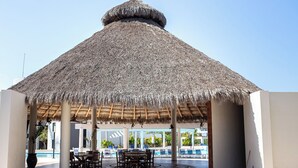 A covered outdoor seating area with a thatched roof, poolside chairs, and a view of the pool and resort buildings. - 3 bedroom penthouse in Bucerias - Ocean view, BBQ and hotel-like amenities (Bucerías)