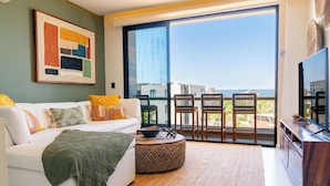 Un salon moderne avec un canapé blanc, une table basse et un téléviseur. Une grande fenêtre offre une vue sur la ville, et un balcon avec des chaises est également présent.