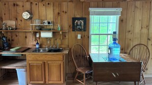 A wooden cabin kitchen with a wooden table and chairs, a sink, and a window with a view of greenery. -  Cozy Off Grid Cabin near the Blueridge Parkway and Appalachian Trail. (Monroe)
