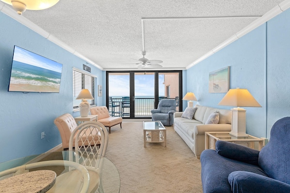 A living room with a view of the ocean, featuring a ceiling fan, a TV, and a seating area with a coffee table. - Sunrise 602 (New Smyrna Beach)