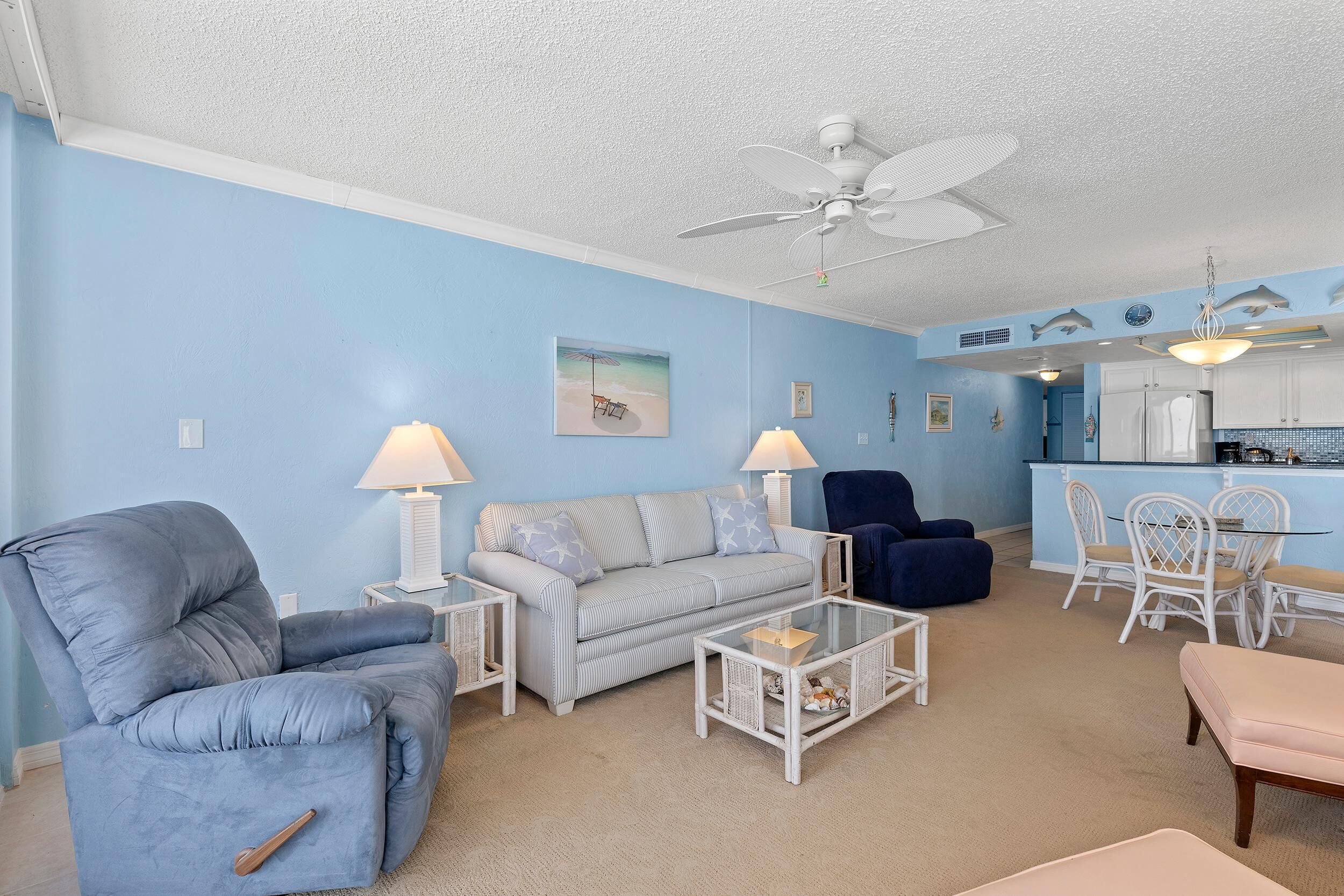A living room with a blue sofa, a blue armchair, a glass coffee table, and a dining area with white chairs.