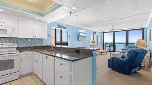 Eine moderne Küche mit weißen Schränken, einer Granit Arbeitsplatte und einer großen Kochinsel. Im Wohnbereich befindet sich ein blauer Sessel und eine Lampe, und es gibt einen Blick aufs Meer durch große Fenster.